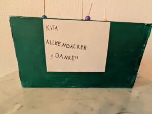 Grün angemalte Rückseite des Miniaturtheaters, auf weißen Papier, welches aufgeklebt ist, steht geschrieben: KITA ALLMENDÄCKER DANKE!