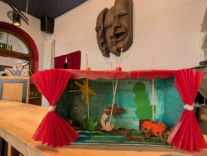 Gastgeschenk der Kita Almendäcker, die beim Kinder- Kunsttag am Forum Theater zu Gast waren. Die Kinder haben ein Miniaturtheater aus einem Schuhkarton gebaut. Diese Miniaturbühne hat einen roten Vorhang und die Wände der Bühne sind grün bemalt, mit einer Sonne links und einem Baum auf der rechten Seite. Von der Decke hängen, mit Holzspießen in einer Art Gleis befestigt, mehrere Tiere, so dass man mit diesem Miniaturtheater ein kleines Figurentheater spielen kann. Am Bühnenboden des Miniaturtheaters befinden sich weitere Ausstattungselemente, u.a. Moos.